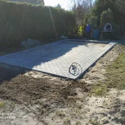 Przygotowany teren pod taras z ułożoną częścią kostki brukowej, widoczne przewody elektryczne, w tle tuje i piec chlebowy obrośnięty zielenią.