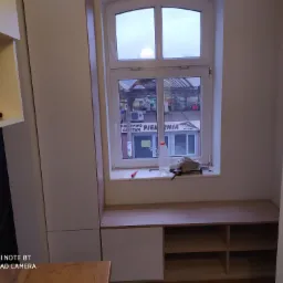 Wnęka okienna z zabudową meblową w trakcie montażu, widoczne narzędzia i materiały na parapecie, częściowo zamontowane szafki z półkami, widok na ulicę przez okno.