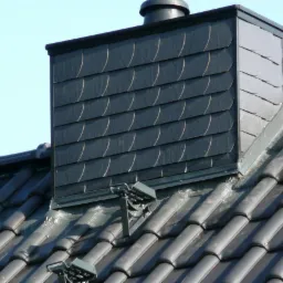 Szczegółowe ujęcie dachu z czarnej dachówki ceramicznej i komina obłożonego łupkiem, widoczne mocowania do zabezpieczenia przeciwśnieżnego.