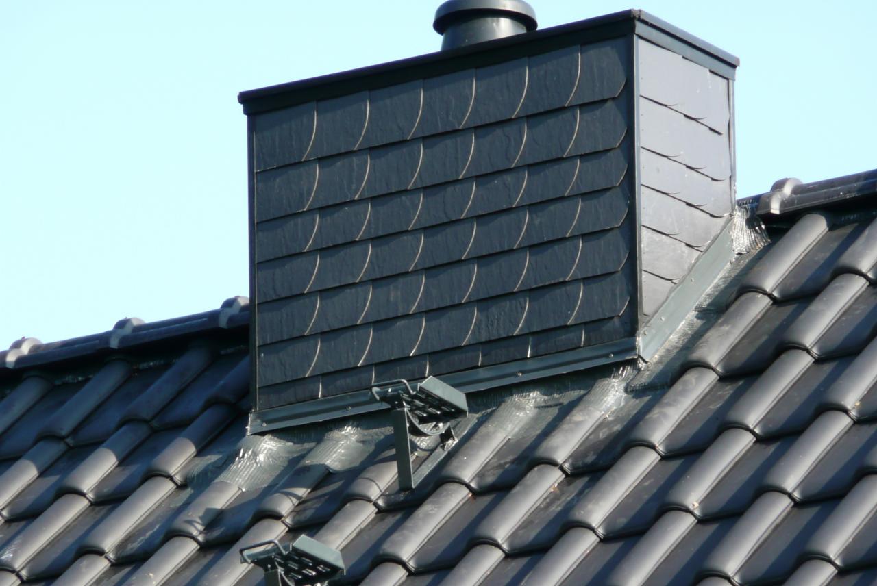 Szczegółowe ujęcie dachu z czarnej dachówki ceramicznej i komina obłożonego łupkiem, widoczne mocowania do zabezpieczenia przeciwśnieżnego.