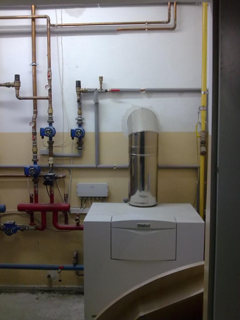 Biały piec Vaillant z metalowym kominem na tle ściany z instalacją hydrauliczną i gazową. Widoczne rury miedziane, zawory i szara skrzynka elektryczna.