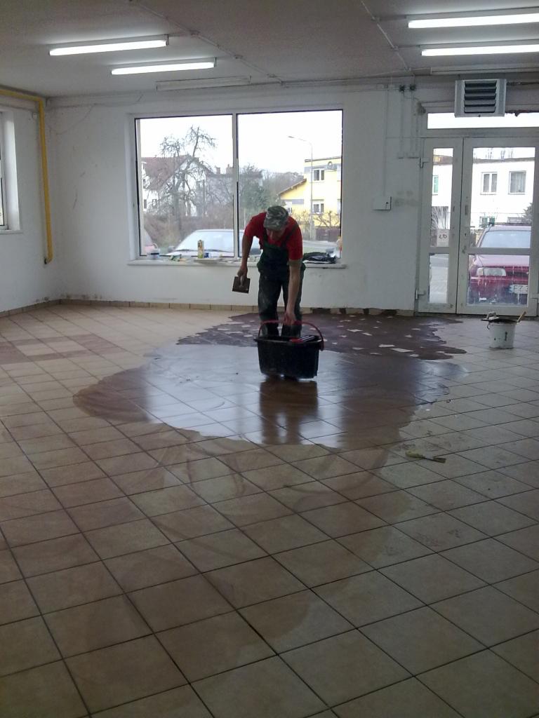 Układanie płytek podłogowych w pomieszczeniu w Jany. Fachowiec w trakcie pracy, widoczny klej na podłodze i narzędzia.