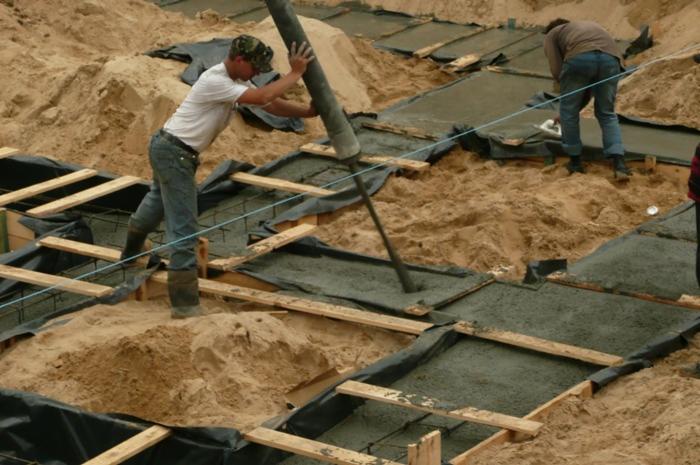 Wylewanie fundamentów: robotnik wlewa beton z rury do szalunków, teren piaszczysty, widoczne drewniane deski i folia. Prace budowlane w toku.