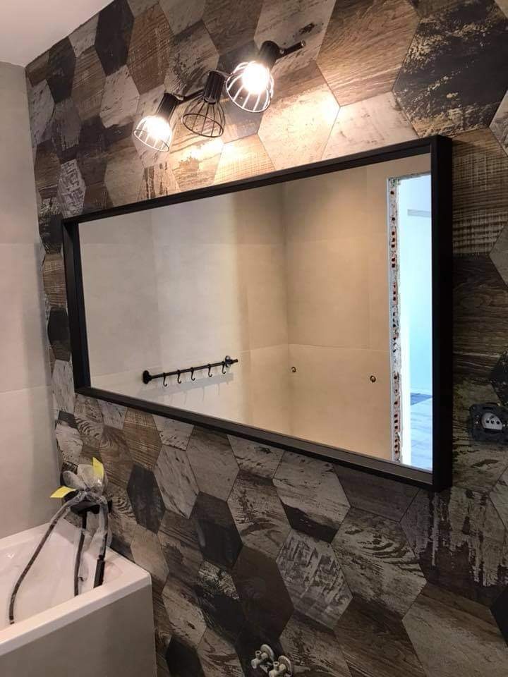 Ukończona łazienka z lustrem w czarnej ramie, heksagonalnymi płytkami imitującymi drewno na ścianie, nowoczesnym oświetleniem i białą umywalką z widocznymi przewodami.