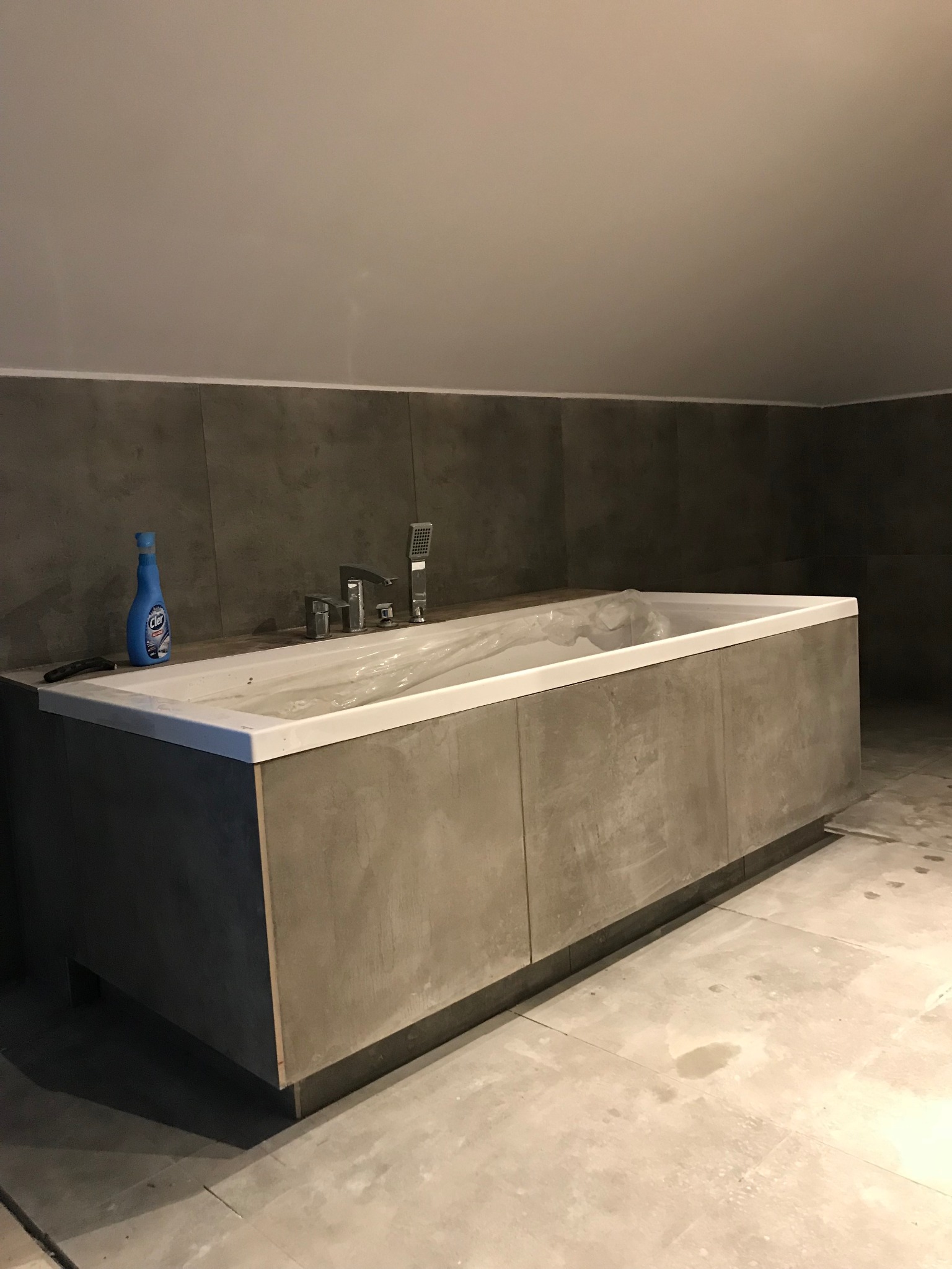 Wanna w trakcie montażu, zabezpieczona folią, z widoczną armaturą i butelką płynu czyszczącego, szare płytki na ścianach, surowy stan wykończenia poddasza.