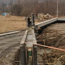 Wykop fundamentów z deskowaniem i świeżo wylanym betonem, widoczne zbrojenie, w tle krajobraz z budynkami i drzewami na wzgórzu.