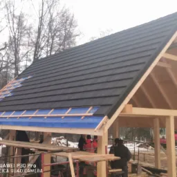 Montaż czarnej blachodachówki na drewnianej konstrukcji dachu, widoczne elementy mocujące i niebieska folia izolacyjna, w tle zimowy krajobraz i pracownicy.