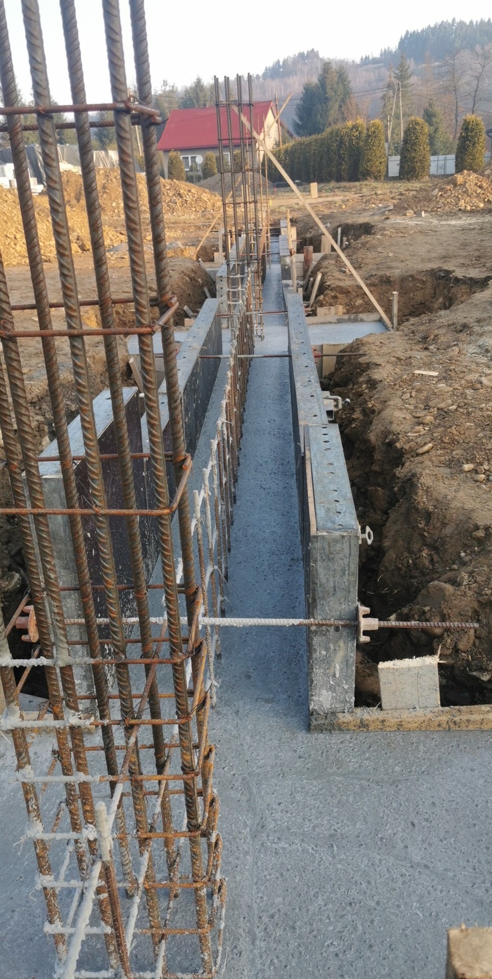 Wylany beton w szalunku fundamentowym z widocznym zbrojeniem pionowym, w tle krajobraz wiejski z domem i drzewami.