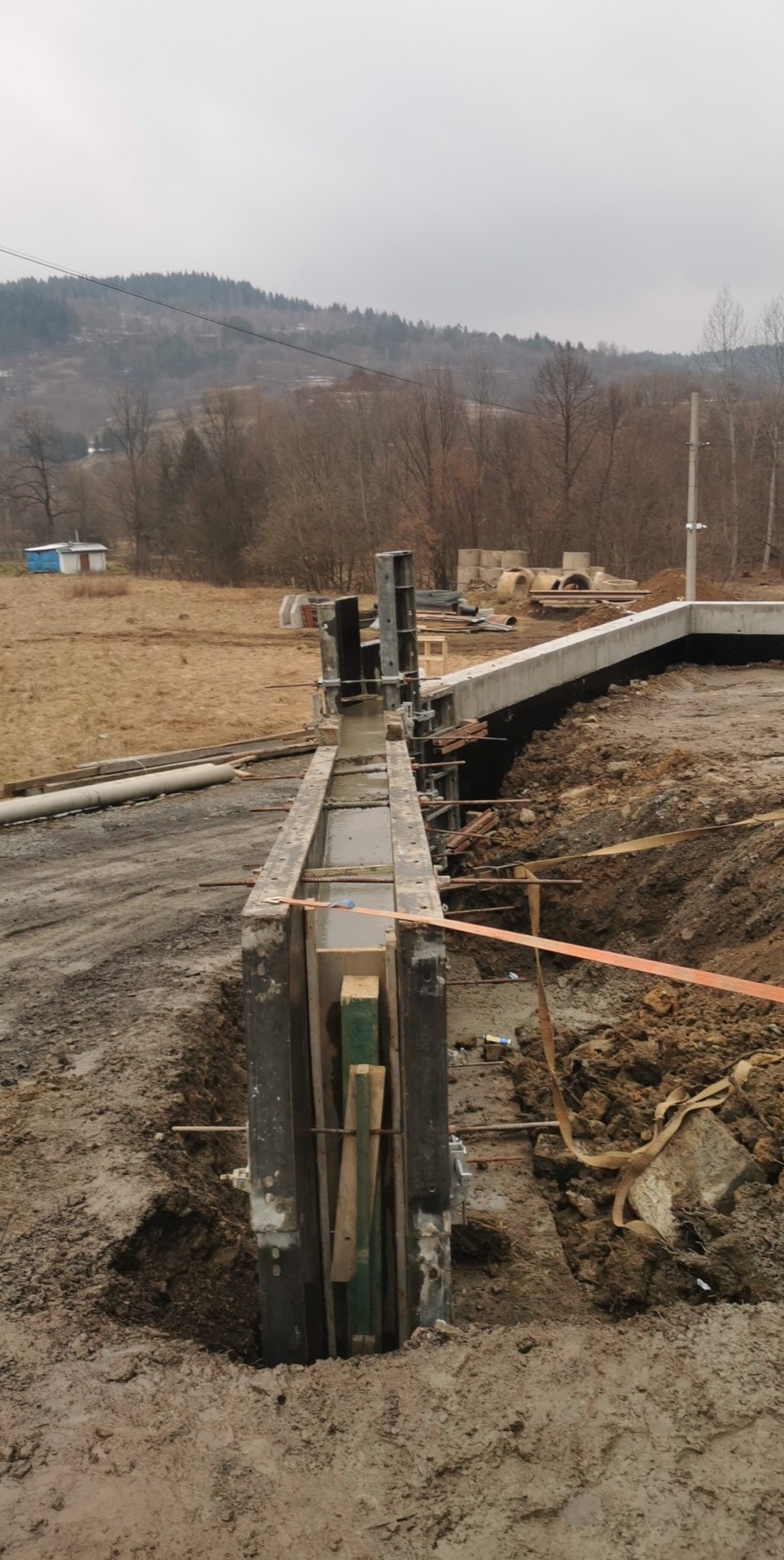 Wykop fundamentów z deskowaniem i świeżo wylanym betonem, widoczne zbrojenie, w tle krajobraz z budynkami i drzewami na wzgórzu.