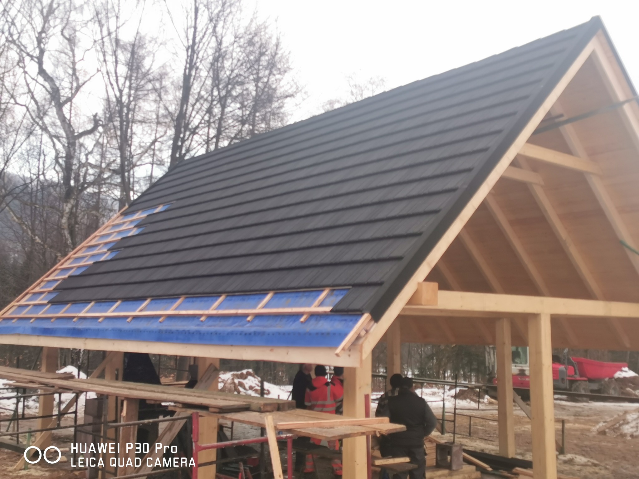 Montaż czarnej blachodachówki na drewnianej konstrukcji dachu, widoczne elementy mocujące i niebieska folia izolacyjna, w tle zimowy krajobraz i pracownicy.