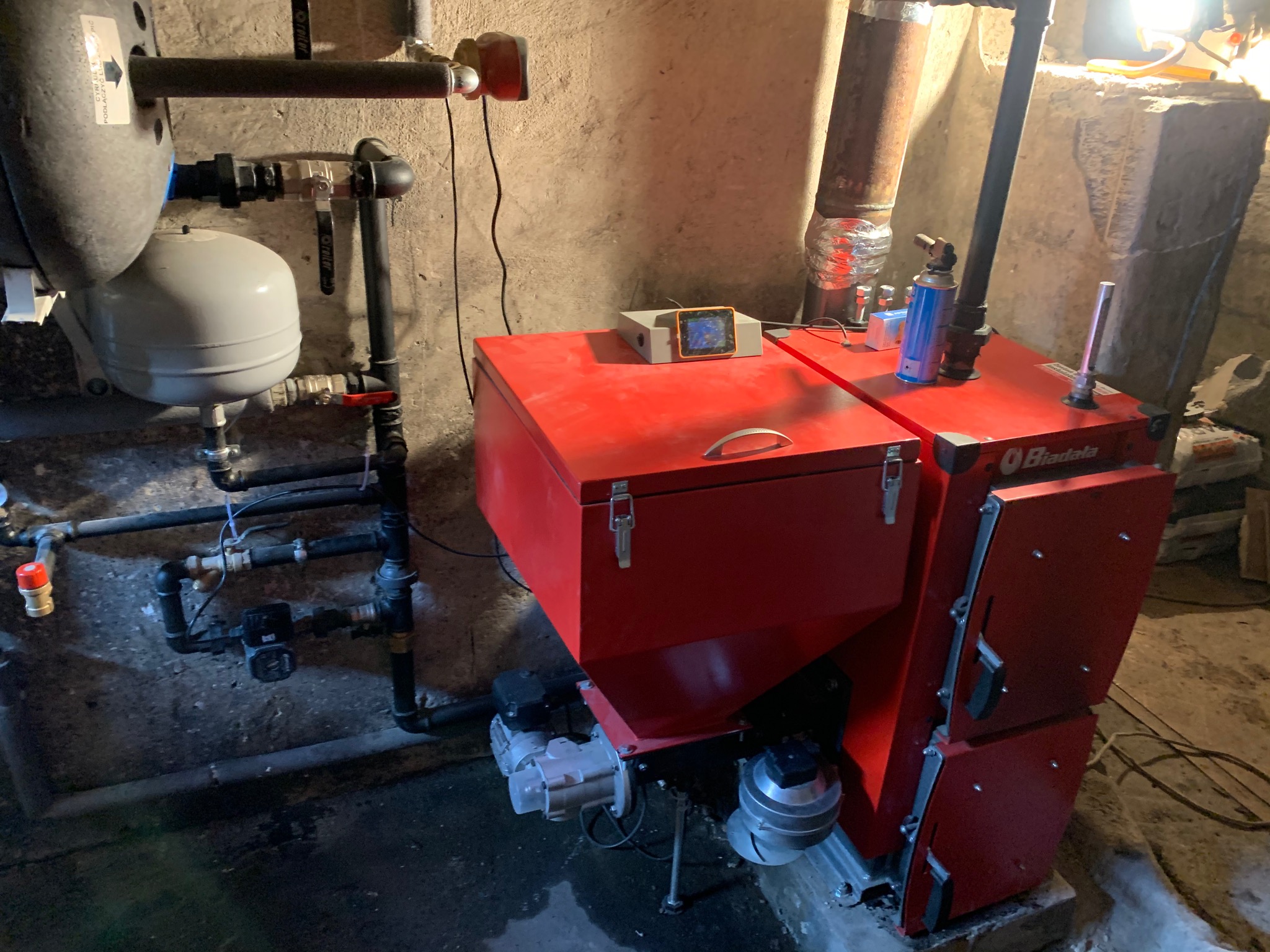 Czerwony piec na paliwo stałe marki Biadala w kotłowni z szarymi ścianami, widoczne rury instalacji grzewczej, sterownik i butla z gazem.
