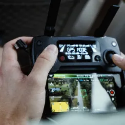 Operator drona trzyma kontroler, ekran pokazuje widok z kamery drona nad zielonym terenem z drogą i jeziorem, tryb GPS aktywny.