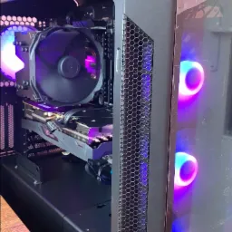 Wnętrze czarnej obudowy komputera stacjonarnego z widocznym chłodzeniem procesora, kartą graficzną Aorus i podświetleniem LED w kolorach fioletu i niebieskiego. Obudowa stoi na drewnianym biurku.