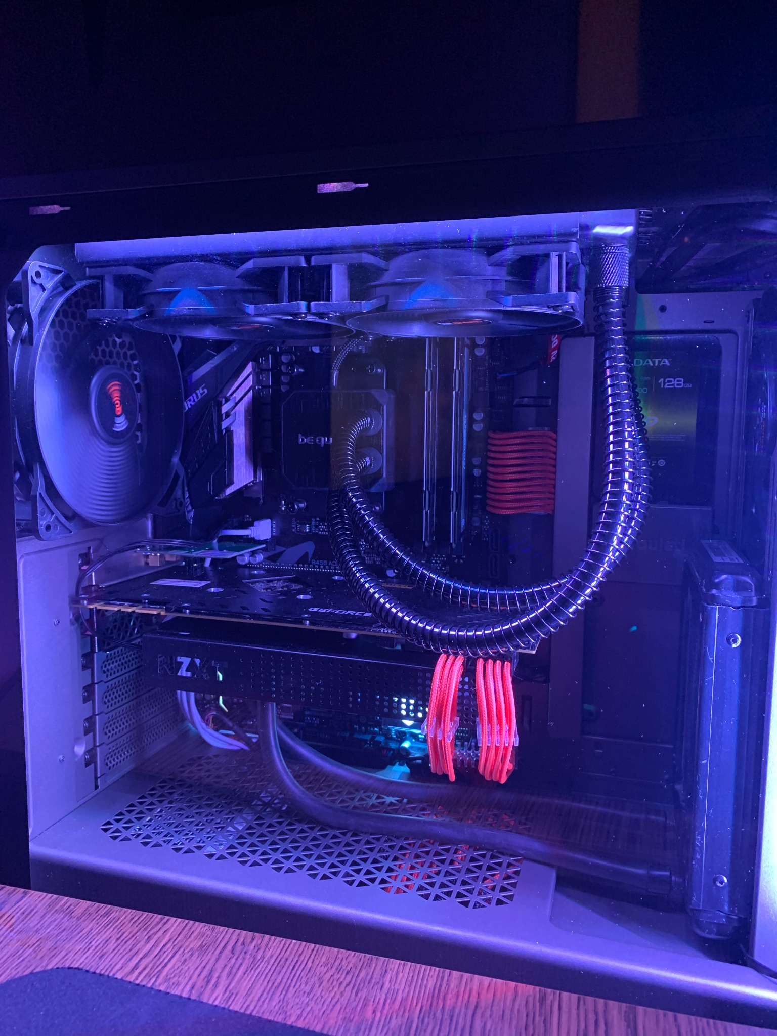 Wnętrze komputera stacjonarnego z widocznym chłodzeniem wodnym, kartą graficzną NZXT, pamięcią RAM, dyskiem SSD, wentylatorami i podświetleniem RGB w odcieniach niebieskiego i czerwonego.