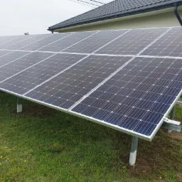 Rząd paneli słonecznych ustawionych na metalowych wspornikach na zielonym trawniku przed budynkiem z ciemnym dachem.