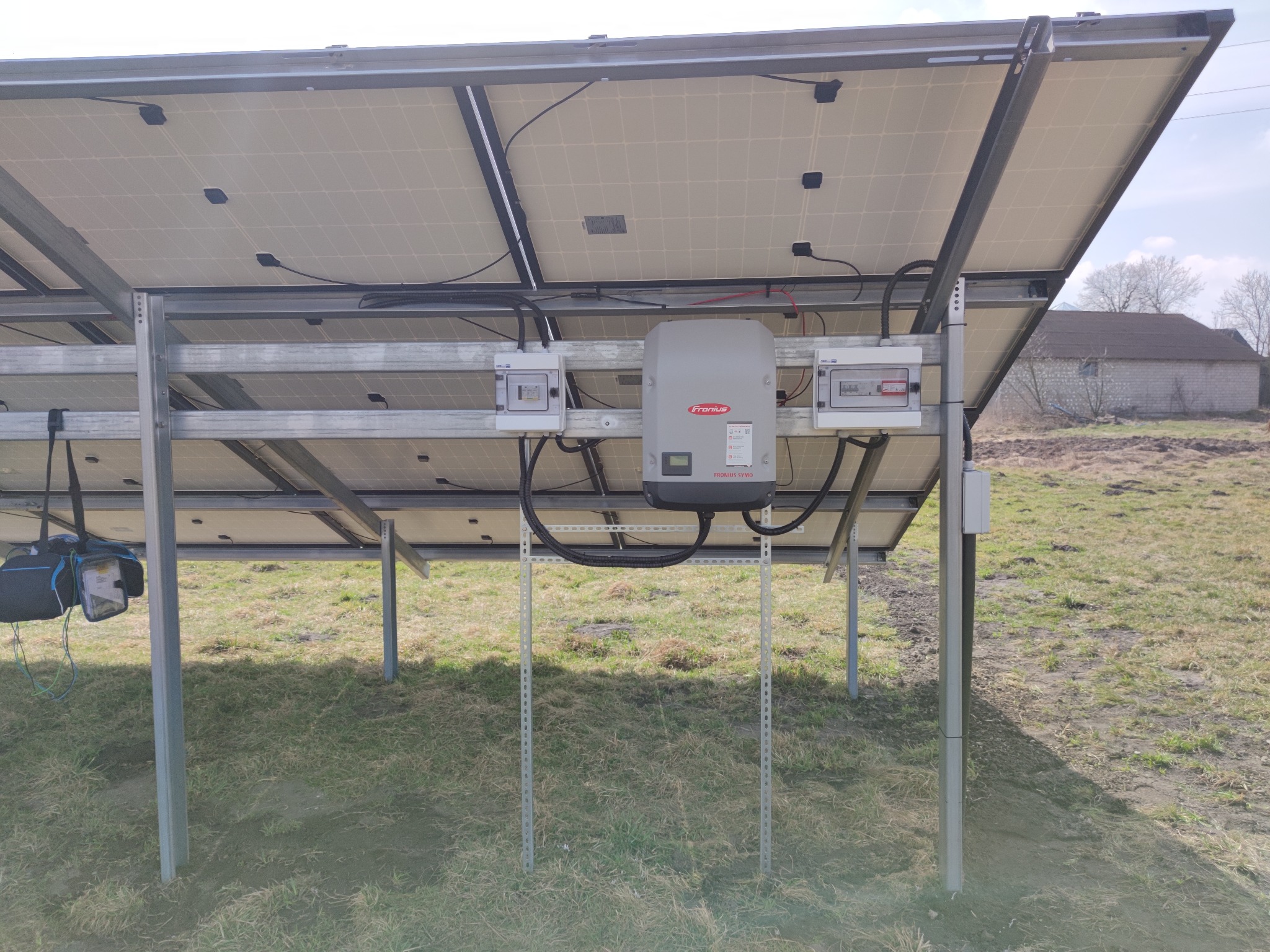 Widok od spodu na panele fotowoltaiczne zamontowane na metalowej konstrukcji, z widocznym szarym inwerterem Fronius i skrzynkami elektrycznymi, na tle zielonej trawy i budynku w oddali.