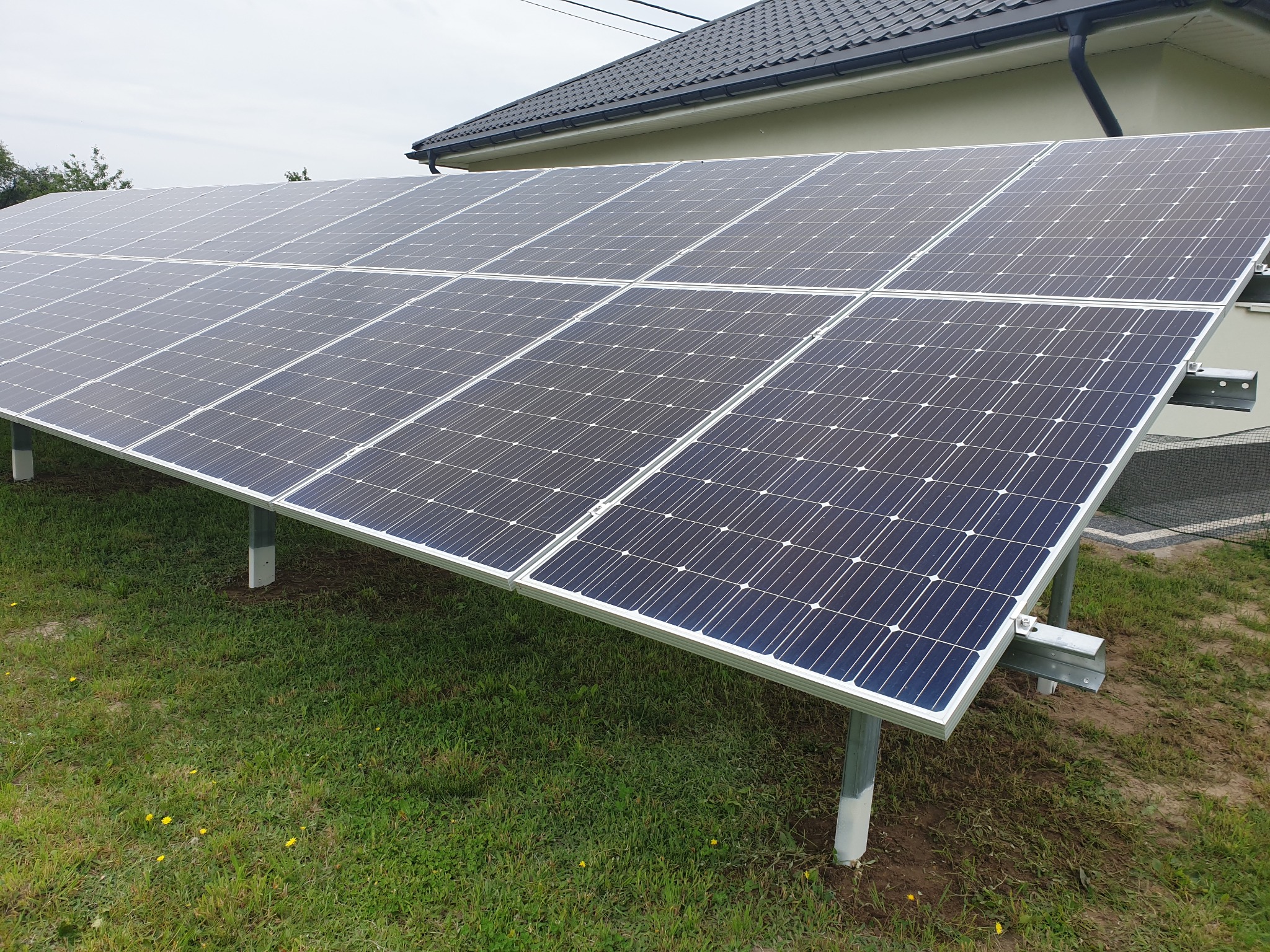 Rząd paneli słonecznych ustawionych na metalowych wspornikach na zielonym trawniku przed budynkiem z ciemnym dachem.