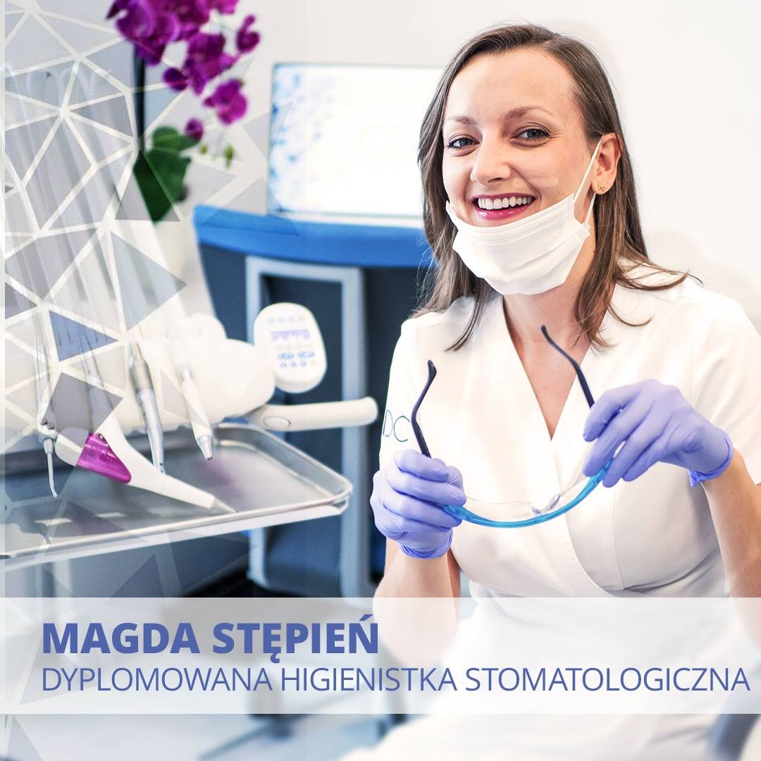 Uśmiechnięta higienistka stomatologiczna w maseczce, trzymająca okulary ochronne w gabinecie z narzędziami dentystycznymi na tacy i fioletowymi kwiatami w tle.