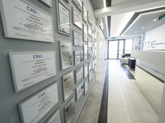 Wnętrze kliniki z wyeksponowanymi certyfikatami i dyplomami na ścianie, widok na recepcję i wejście.