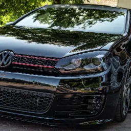 Błyszczący, czarny Volkswagen Golf GTI z czerwonymi akcentami na grillu, nisko zawieszony, zaparkowany na brukowanym podłożu, z odbiciami drzew na karoserii.