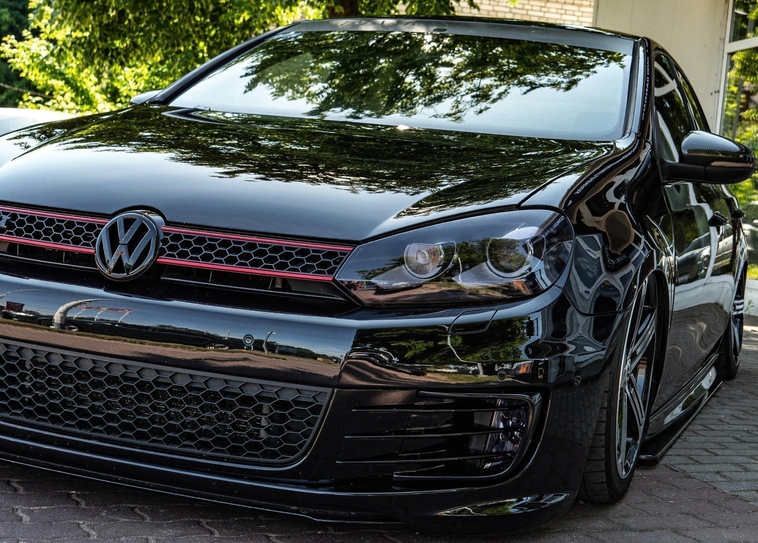 Błyszczący, czarny Volkswagen Golf GTI z czerwonymi akcentami na grillu, nisko zawieszony, zaparkowany na brukowanym podłożu, z odbiciami drzew na karoserii.