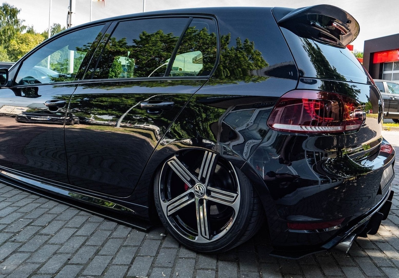 Czarny samochód marki Volkswagen Golf z obniżonym zawieszeniem i felgami aluminiowymi, zaparkowany na kostce brukowej, z widocznym odbiciem otoczenia w lakierze.