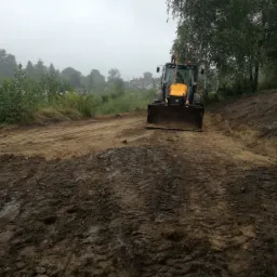 Żółta koparko-ładowarka na skraju wykopu w mglisty dzień, widoczne ślady opon w błocie i brzeg jeziora w tle.