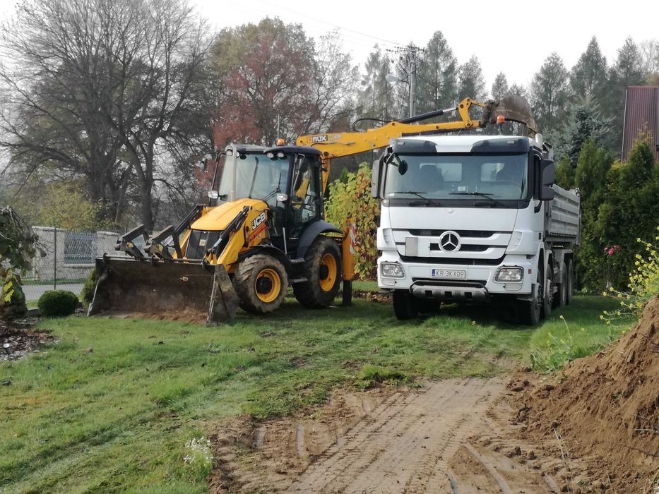 Żółto-czarna koparko-ładowarka JCB i biała wywrotka Mercedes-Benz na trawiastym terenie z drzewami w tle, ślady opon na ziemi.