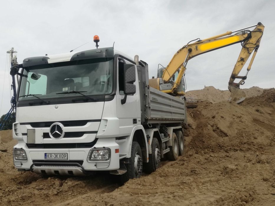 Biała ciężarówka Mercedes z podniesioną skrzynią ładunkową, na której stoi żółta koparka nabierająca ziemię z hałdy. W tle widoczna konstrukcja wiertnicza. Teren wokół pojazdów błotnisty.