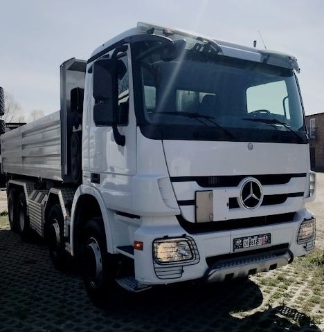 Biała ciężarówka wywrotka marki Mercedes, zaparkowana na betonowej kostce brukowej, z widoczną tablicą rejestracyjną i logo firmy.