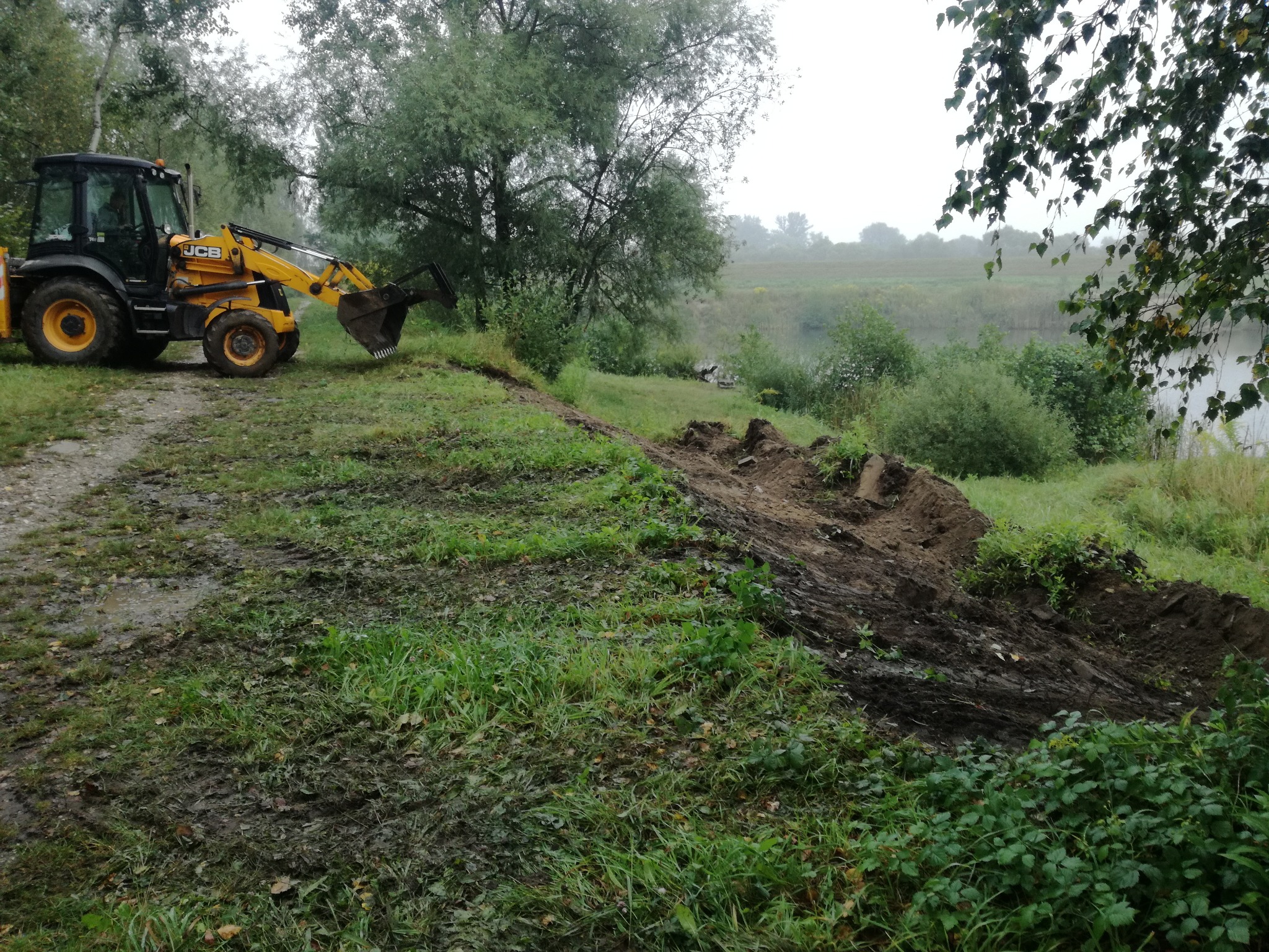 Żółta koparko-ładowarka JCB na skraju terenu z wykopaną ziemią, prawdopodobnie w trakcie niwelacji terenu, w tle widoczny zbiornik wodny i zarośla, pochmurny dzień.