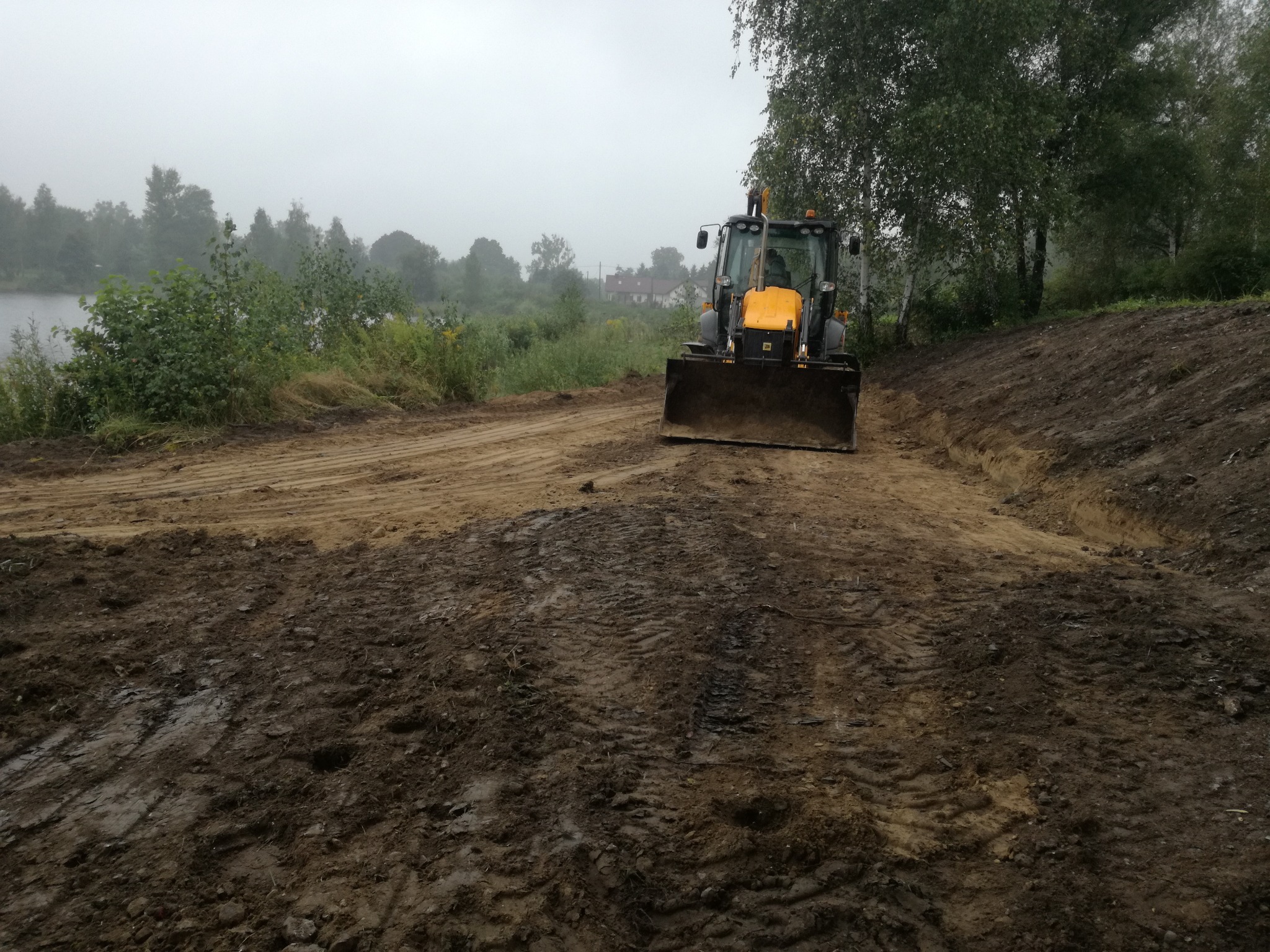 Żółta koparko-ładowarka na skraju wykopu w mglisty dzień, widoczne ślady opon w błocie i brzeg jeziora w tle.