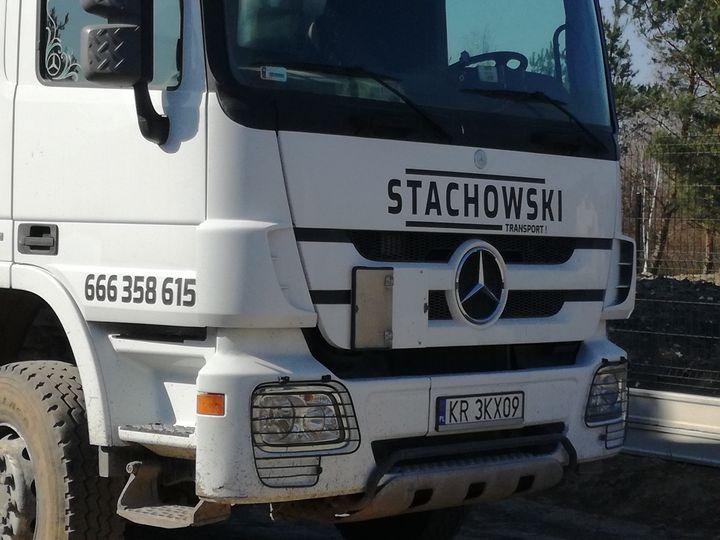 Biały samochód ciężarowy marki Mercedes z numerem telefonu i nazwą firmy transportowej STACHOWSKI TRANSPORT na kabinie, widok z przodu, tablica rejestracyjna KR 3KX09.