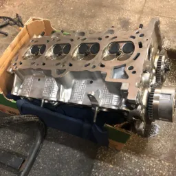 Głowica silnika spalinowego z widocznymi zaworami, umieszczona w kartonowym pudełku na brudnej posadzce warsztatowej, obok widoczne przewody i metalowa konstrukcja w tle.