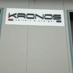 Elewacyjna tablica z nazwą firmy KRONOS battery & charger, czarne litery z czerwonym i niebieskim akcentem.