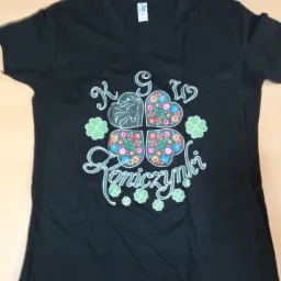 Czarna koszulka z nadrukiem koniczyn, kwiatów i napisu 'Koniczynki' w stylu ludowym, leżąca na jasnym blacie.