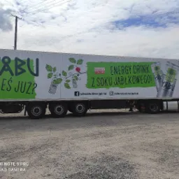 Ciężarówka z reklamą napoju energetycznego YABU z sokiem jabłkowym, z zielonymi elementami graficznymi i hasłem 'Piłeś już?', zaparkowana na szutrowym placu.