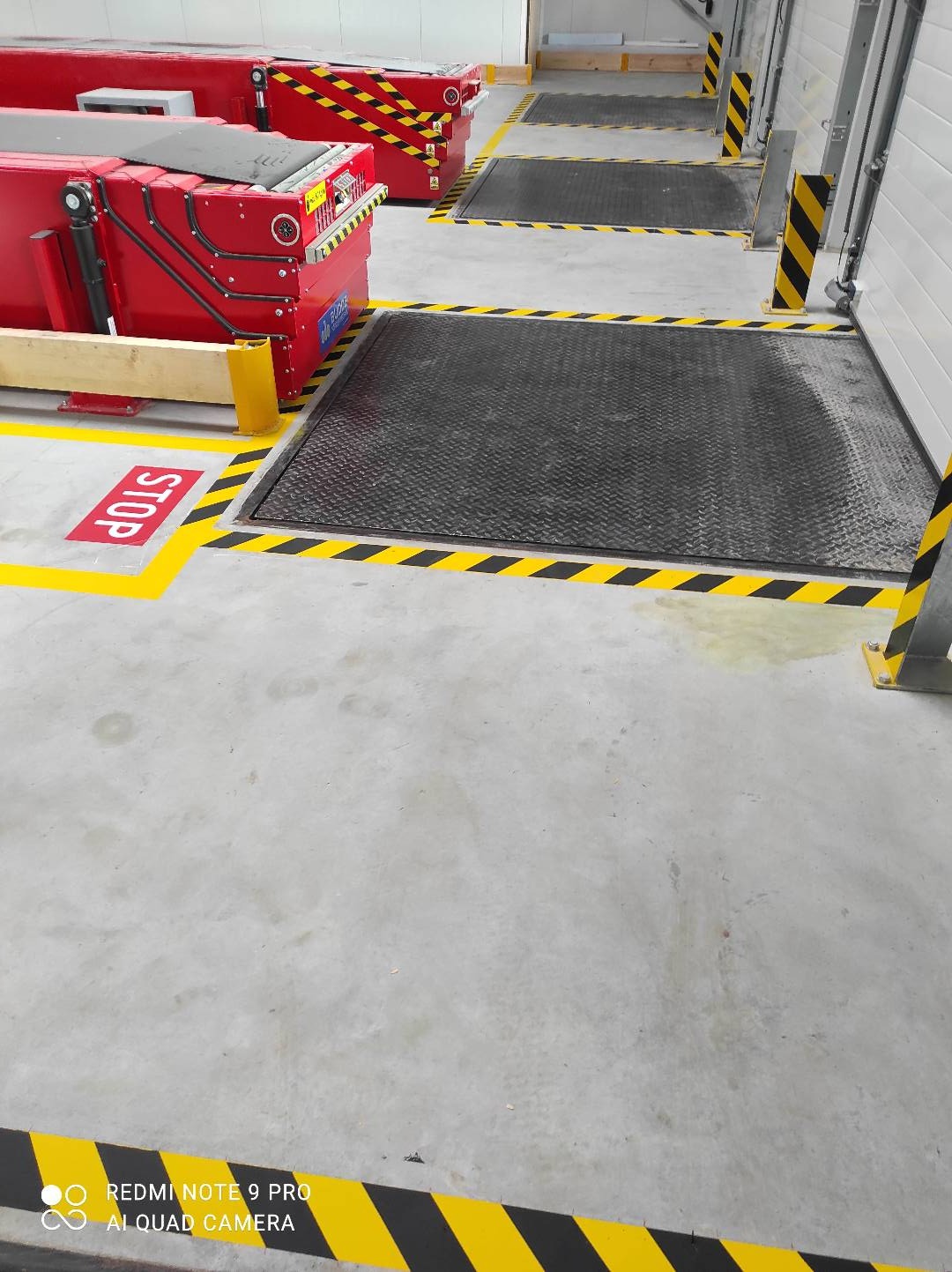 Oznakowanie poziome w hali magazynowej: czerwone skrzynie z żółto-czarnymi pasami, metalowe kraty w podłodze otoczone żółto-czarnymi pasami, napis STOP na podłodze.