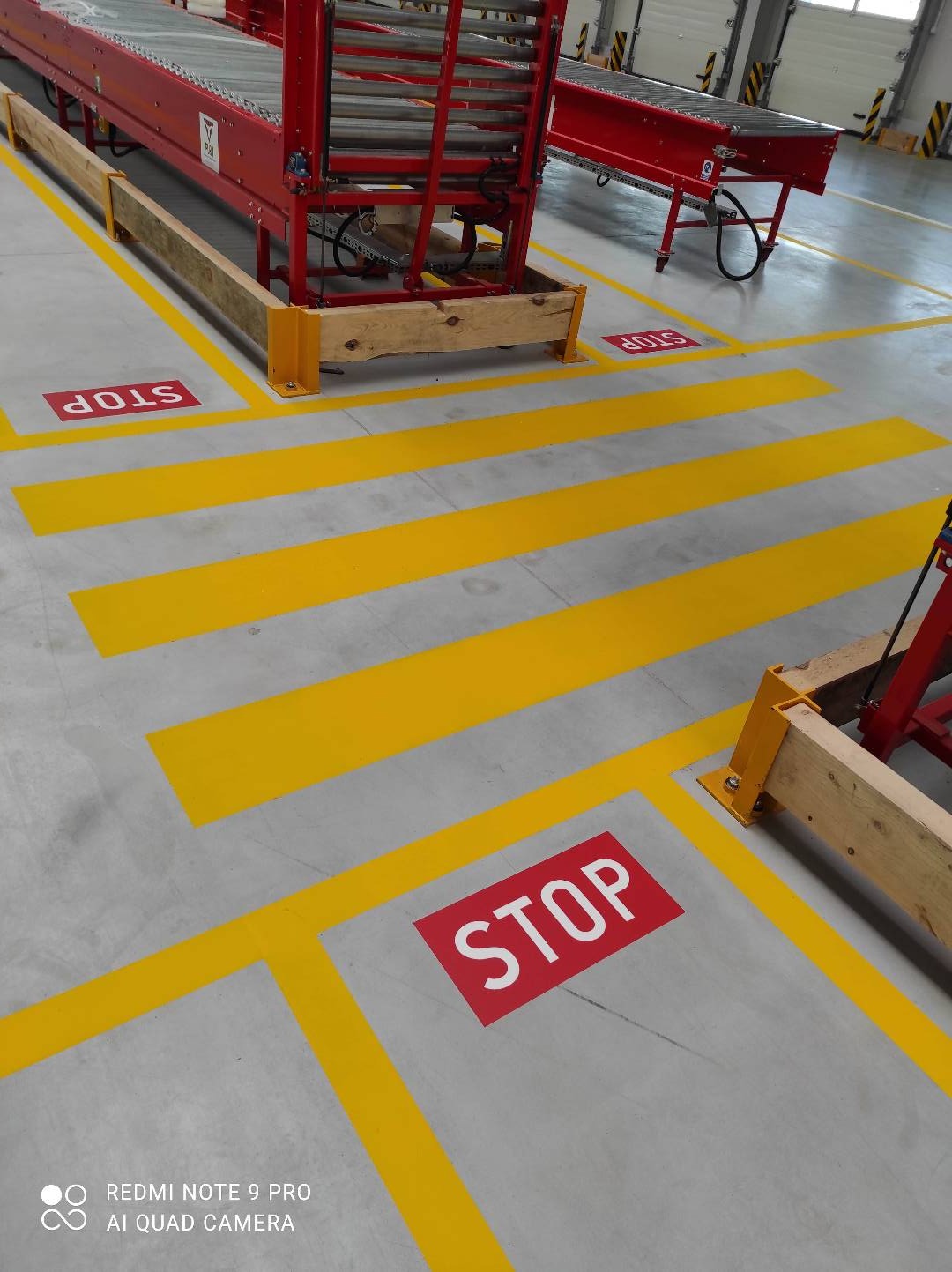 Żółte pasy i czerwone znaki STOP wyznaczające strefy na posadzce hali produkcyjnej, widoczne fragmenty czerwonych konstrukcji metalowych.