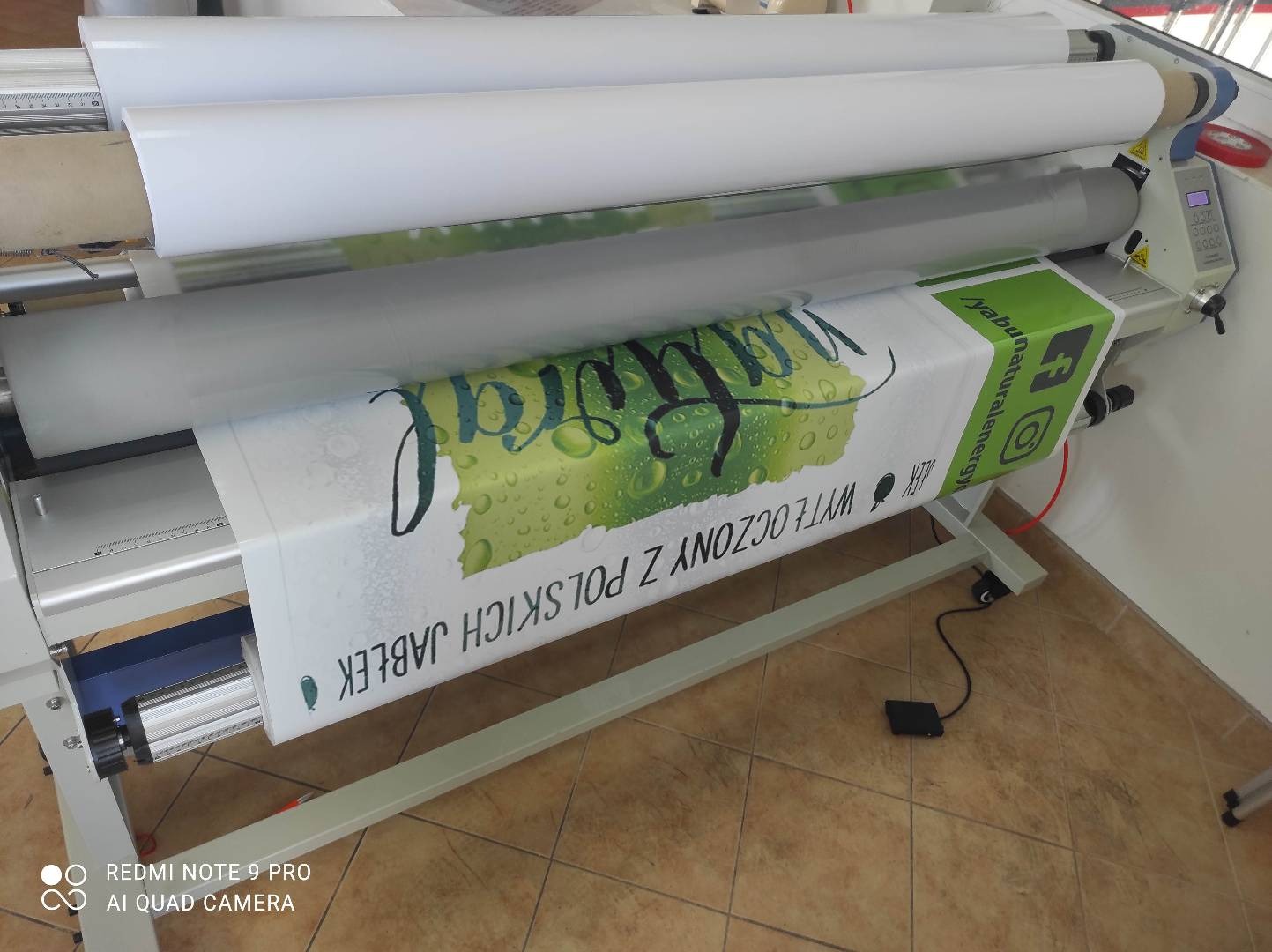 Proces laminowania wydruku wielkoformatowego z motywem jabłek i napisem 'Wy tłoczony z polskich jabłek' na maszynie drukarskiej.