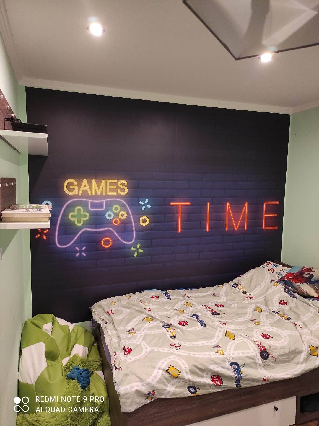 Pokój dziecięcy z czarną ścianą ozdobioną neonowym napisem 'GAMES TIME' i motywem kontrolera do gier, łóżko przykryte pościelą z motywem samochodów i dróg, obok zielony puf.