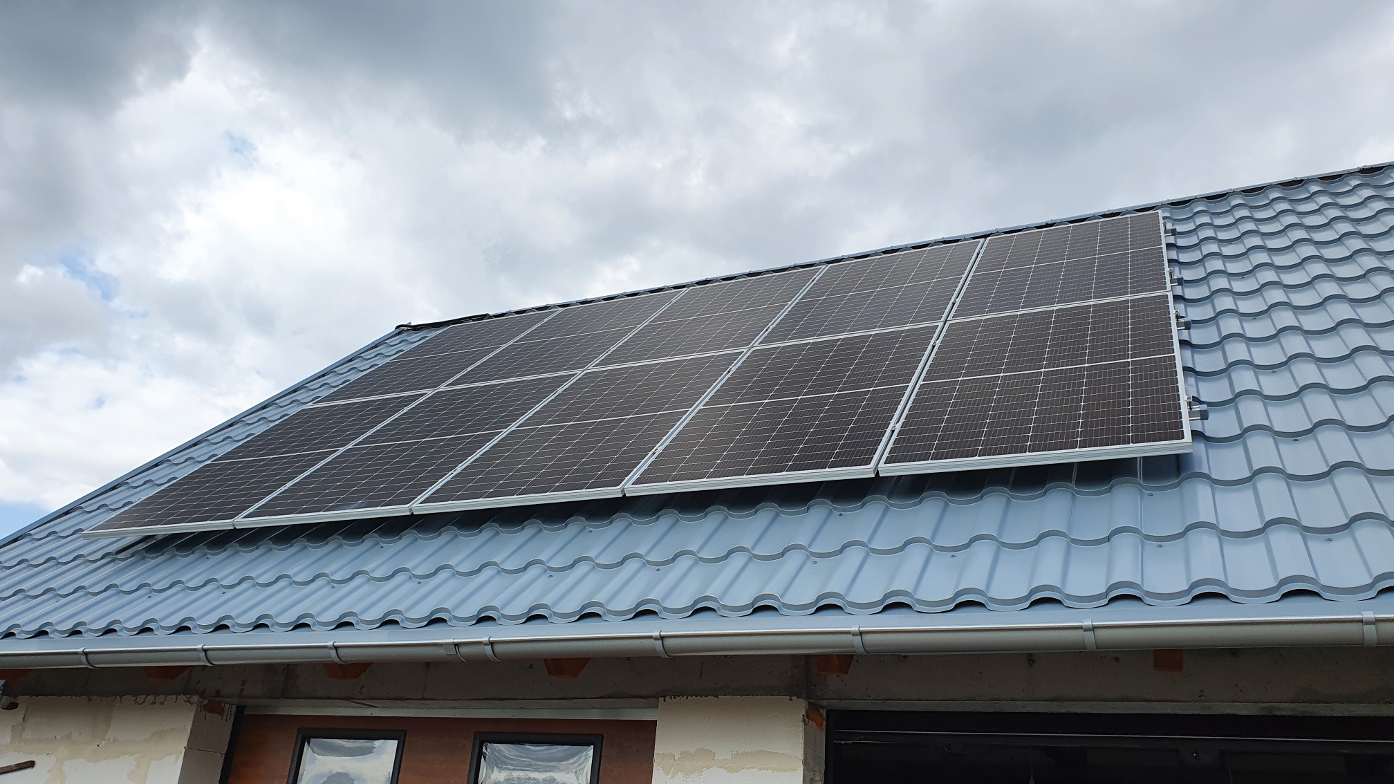Panele słoneczne zamontowane na nowym, błękitnym dachu z blachodachówki, widoczne z dołu na tle pochmurnego nieba. Częściowo otynkowana elewacja budynku w stanie surowym.