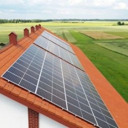 Synergio Spółka Akcyjna - Panele słoneczne na dachu domu z czerwoną dachówką, widok z góry na zielone pola w tle.