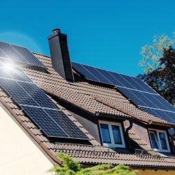 Synergio Spółka Akcyjna - Instalacja fotowoltaiczna na brązowym dachu domu jednorodzinnego z kremową elewacją, widoczne okna z białymi ramami i czarny komin, jasne słońce odbija się od paneli.