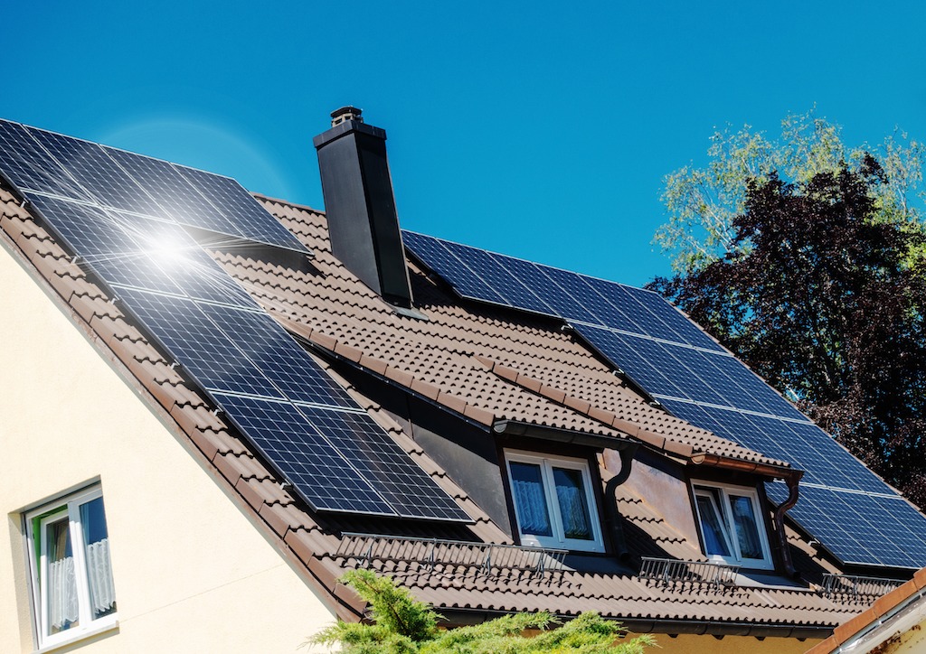 Instalacja fotowoltaiczna na brązowym dachu domu jednorodzinnego z kremową elewacją, widoczne okna z białymi ramami i czarny komin, jasne słońce odbija się od paneli.