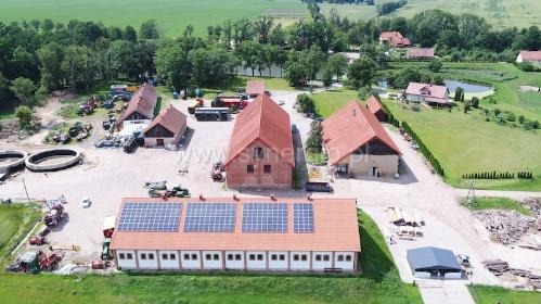 Widok z lotu ptaka na budynek gospodarczy z panelami słonecznymi na dachu, otoczony zabudowaniami gospodarczymi i zielonymi polami.