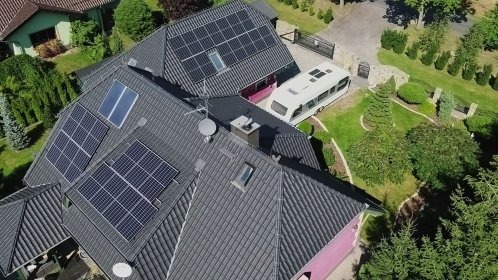 Widok z góry na dom z ciemnym dachem, na którym zamontowano panele słoneczne; na podjeździe stoi biały bus.