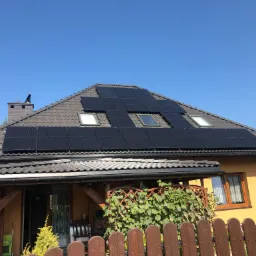 Żółty dom jednorodzinny z brązową dachówką, na której zamontowane są czarne panele fotowoltaiczne oraz okna dachowe. Widoczny komin z daszkiem i drewniany płot.