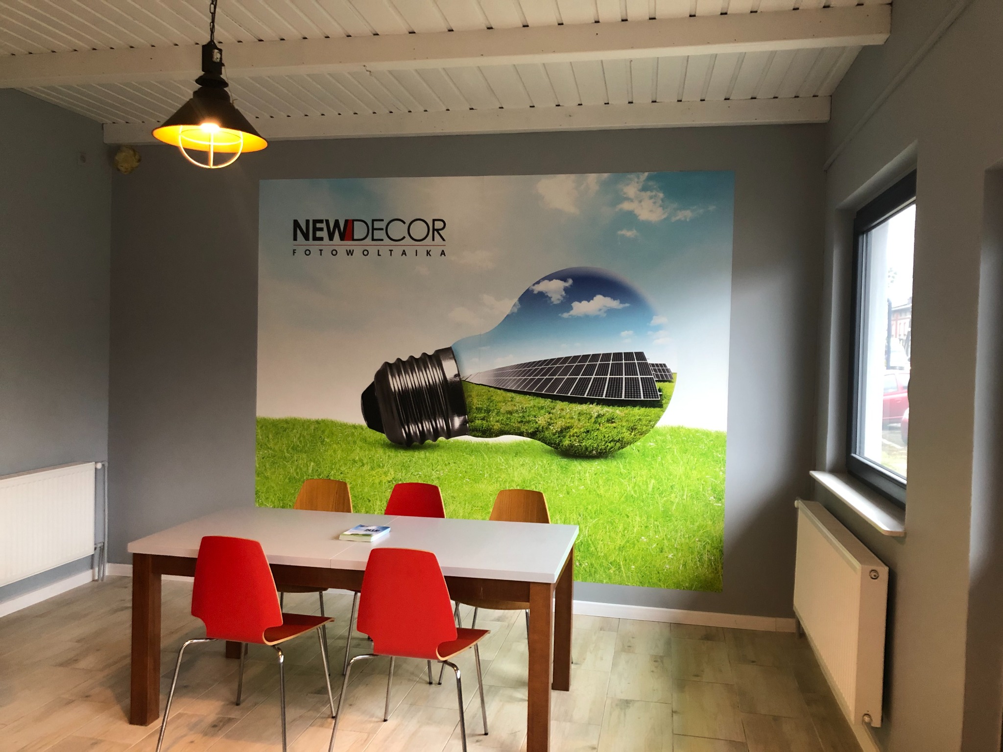 Wnętrze biura z plakatem fotowoltaiki przedstawiającym żarówkę wypełnioną panelami słonecznymi na tle zielonej trawy i błękitnego nieba, nad białym stołem z czerwonymi i brązowymi krzesłami...