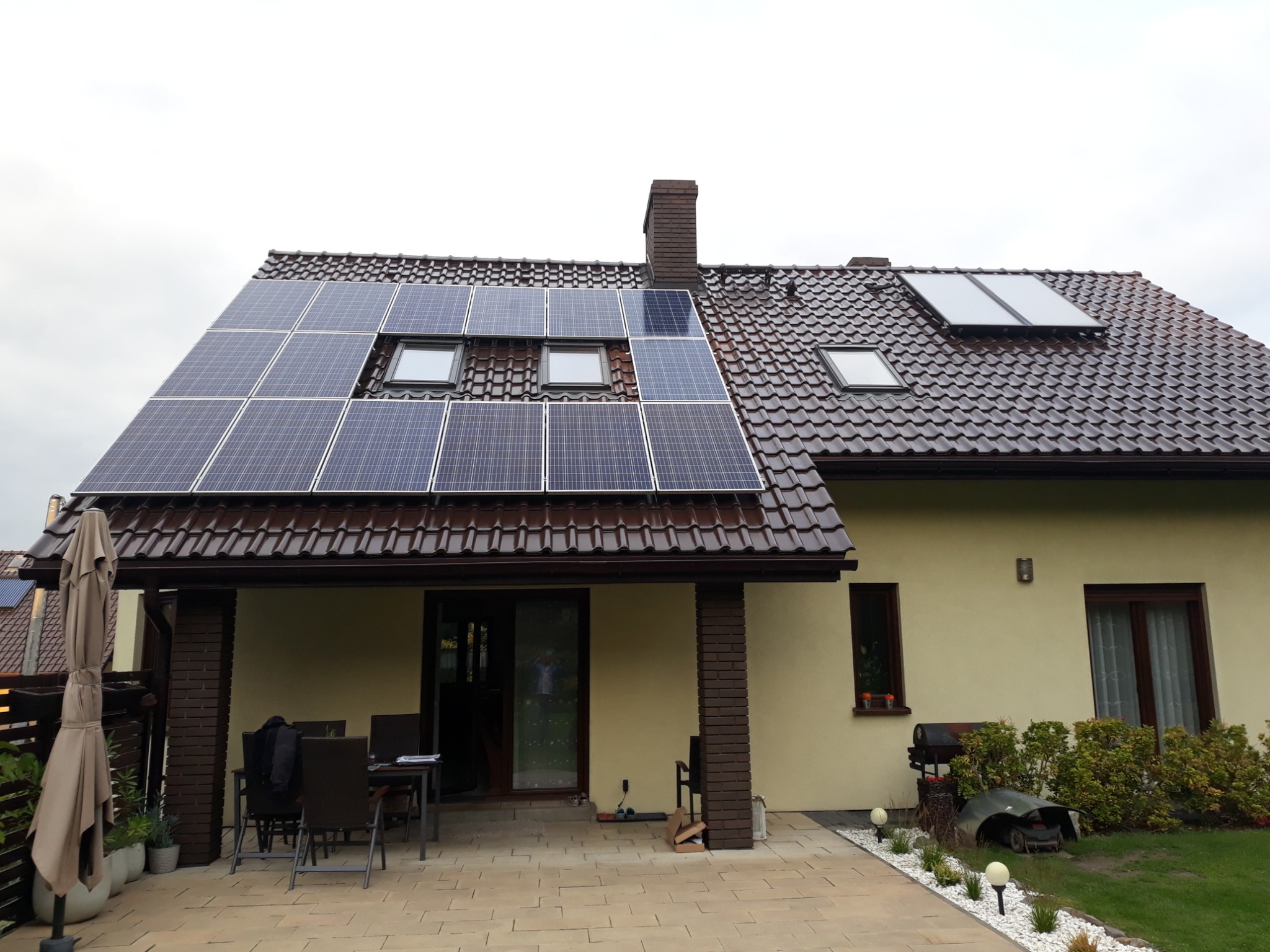 Dom jednorodzinny z brązowym dachem, na którym zamontowano panele fotowoltaiczne, widoczne także okna dachowe i komin.